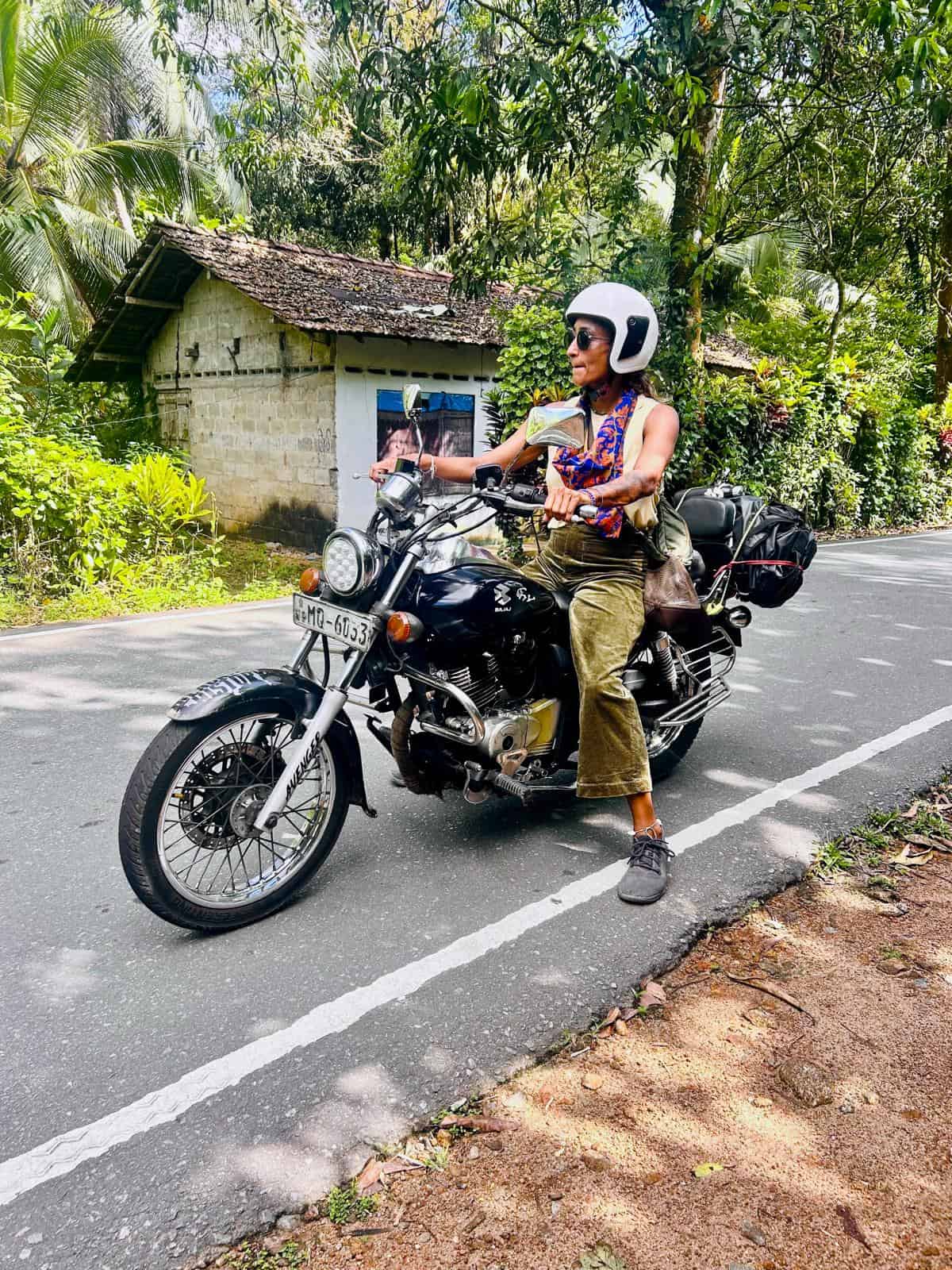 Woman riding motorcycle with helmet on rural road surrounded by greenery.